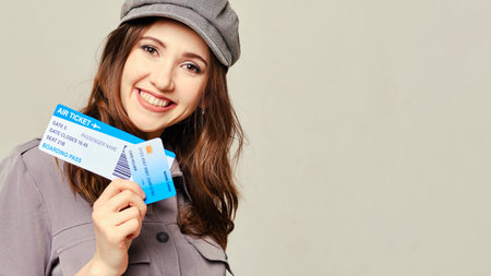 A girl in a gray dress looks at the camera and holds plane tickets with a credit card. Copy space....