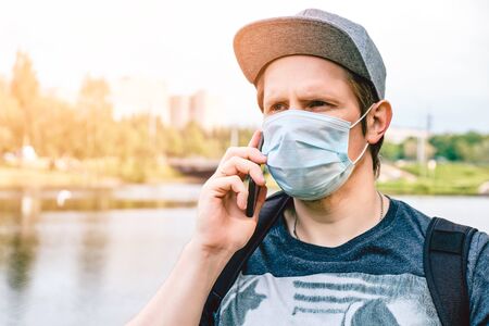 Caucasian man with a medical mask and a cap walks down the street in a park during a pandemic and speaks on a smartphone.の写真素材