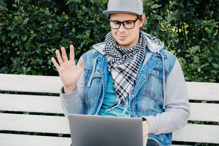 A Caucasian man sits on a bench and communicates via video chat on a laptop