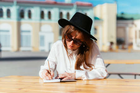 Caucasian girl in a hat and sunglasses smiling while sitting in a cafe in the summer and makes notes in a notebook.の写真素材