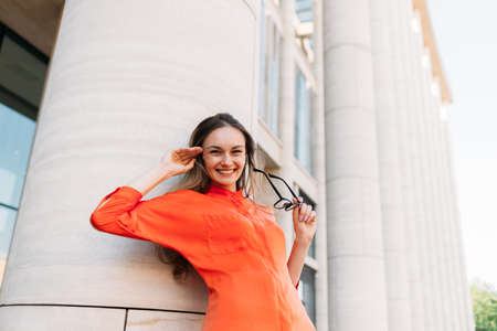 Caucasian girl joyful holds glasses in her hands and leans on a modern building.の写真素材