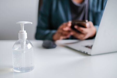 An alcohol gel bottle place on a white office desk is used to clean hands for protective COVID-19 with an office worker working nearby and a notebook. Concept work from homeの写真素材