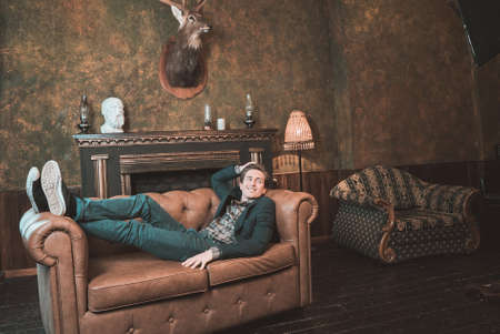 The man is lying on a brown leather sofa near the fireplace