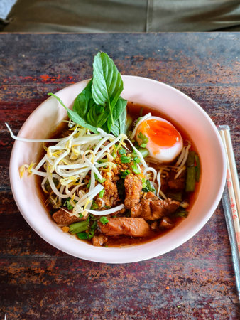 A steaming bowl of Thai beef noodle soup featuring tender beef slices, rice noodles, bean sprouts, green onions, crispy garlic, basil, and a runny soft-boiled egg in rich dark broth.の写真素材