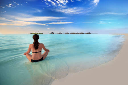 Young woman exercising yoga in turquoise lagoon with overwater bungalows around tropical islandの写真素材