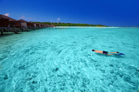 Young man snorkeling in tropical island with sandy beach, palm trees, overwater bungalows and turquoise clear waterの写真素材