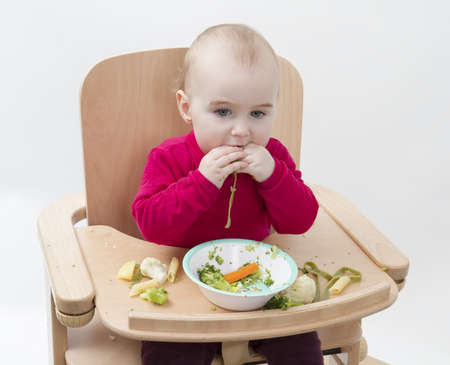 young child in red shirt eating vegetables in wooden chair の写真素材