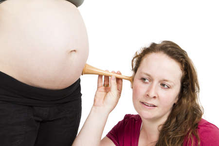 young midwife listening at human belly  pregnant woman standing while midwife is sitting in front ...
