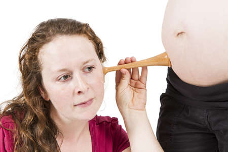 young midwife listening at human belly  pregnant woman standing while midwife is sitting in front  horizontal picture isolated on whiteの写真素材