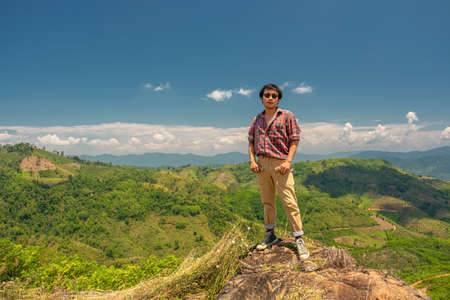 Asian man standing on a rock against a backdrop of mountains and sky. A 35-year-old man wearing a striped shirt and brown trousers with sunglasses stands on a rockの写真素材