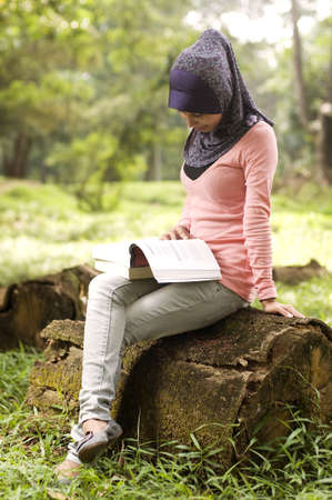 Beautiful young muslim lady sit on the stump reading isolated green parkの写真素材