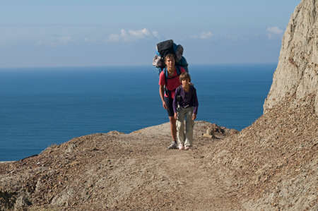 The woman and her daughter moves with backpacks along the rock. Tourists are situated against the bue sea and sky background.の写真素材