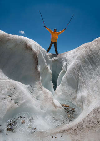 Woman dressed in bright orange wind jacket on the glacier top. Tourist puts up hands with sticks. Scene against blue sky background.の写真素材