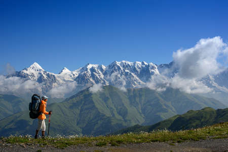 Backpacker with sticks goes along mountain range. Mountain with snow and blue sky are in the...