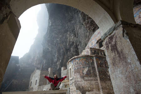Woman dressed in red clothes meditates in Sumela Monastery Turkey. Foggy weather enhances spiritual discipline effect.の写真素材