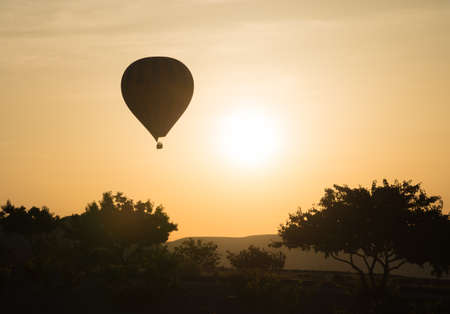 Hot-air balloon flying at sunrise. Trees silhouettes in foreground. Gold sky in background.の写真素材
