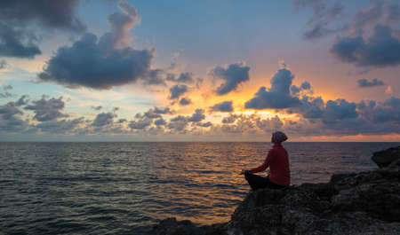 Woman sits as yogi on rock. Beautiful sunset over sea in background.の写真素材