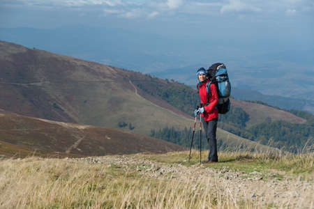 Tourist dressed in bright red windbreaker with sticks and big backpack in mountains.の写真素材
