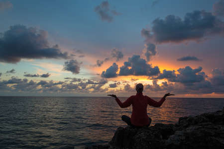 Woman (or man) sits as yogi with outstretched hands and contemplates beautiful sunset over sea.の写真素材