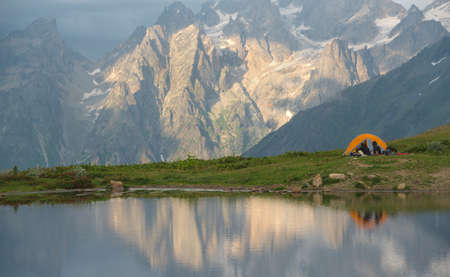 Tourists with orange tent near mountain lake. Rocks with snow and glacier in background.の写真素材