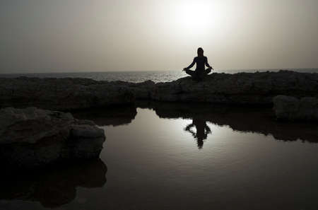 Woman sits as yogi on rock and meditates. Sea in background. Woman's silhouette reflects in water.の写真素材