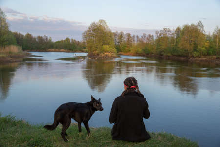 Girl with black dog is sitting on green grass meadow under river. Sky, trees and island in background.の写真素材