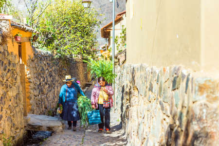 OLLANTAYTAMBO, PERU - JUNE 26, 2019: Peruvian women on a city street carry large armfuls of grassのeditorial素材