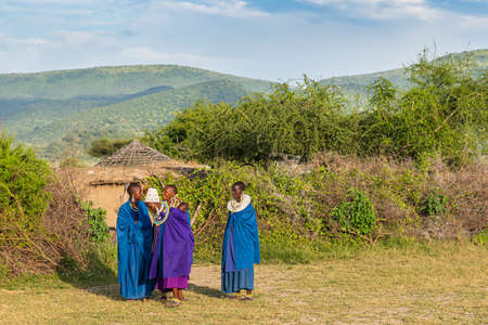 TANZANIA, AFRICA-FEBRUARY 15, 2020: Four  Massai Women and one Baby with traditional Dresses stay smiling at Sunset together. In the Background a part of a Hut, Plants and Mountainのeditorial素材