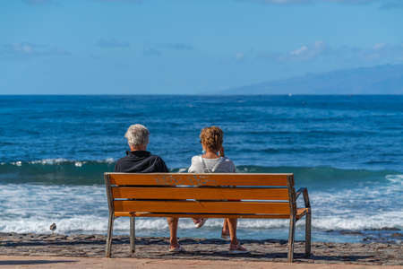 Back view of man and woman sitting on bench and looking to the ocean, outdoors background. Back view of people panoramaの写真素材