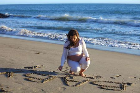 Pregnant woman at the beach writing  baby in the sand near the oceanの写真素材
