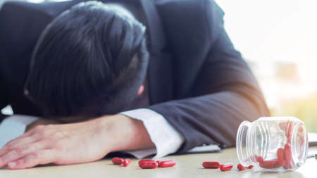 Asian businessman sleeps on his desk in fatigue and scattered painkillers on the tableの写真素材