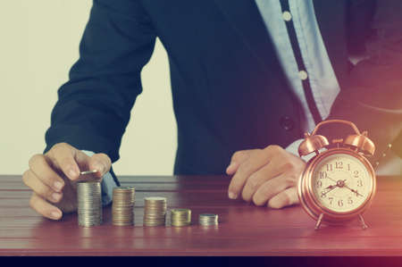 business man put money on pile of coins with alarm clock on working table, concept in time to successの写真素材