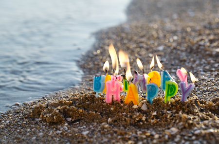 Colourful birthday candles spelling out the alphabet letters - Happy Birthday - standing upright in beach sand burning on a seashore at the edge of the seaの写真素材