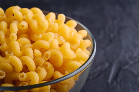 Glass bowl with cavatappi uncooked golden wheat curly pasta on textured dark black background, angle view macroの写真素材