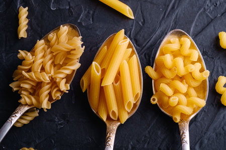 Three cutlery spoons with variety of uncooked golden wheat pasta on dark black textured background, top view macroの写真素材