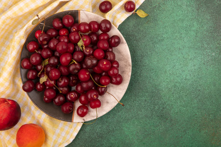 top view of cherries in plate with peach and apricot on plaid cloth on green background with copy...
