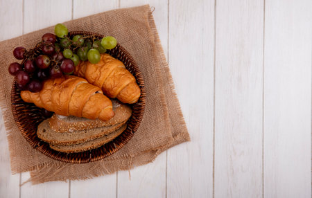 top view of breads as croissant and seeded brown cob bread slices with grape in basket on sackcloth on wooden background with copy spaceの写真素材