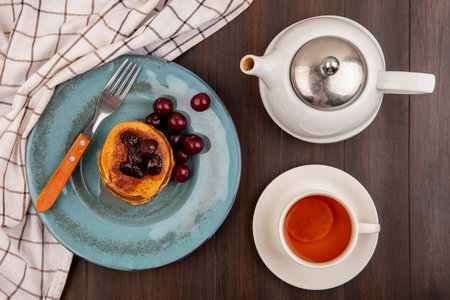 top view of breakfast set with pancake and cherries and fork in plate on plaid cloth and cup of tea with teapot on wooden backgroundの写真素材