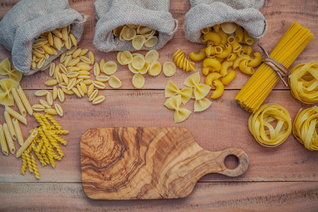 Italian foods concept and menu design . Various kind of Pasta Elbow Macaroni ,Farfalle ,Rigatoni ,gnocco Sardo in hemp sack bags set up on brown wooden background with cutting board for copy space .の写真素材