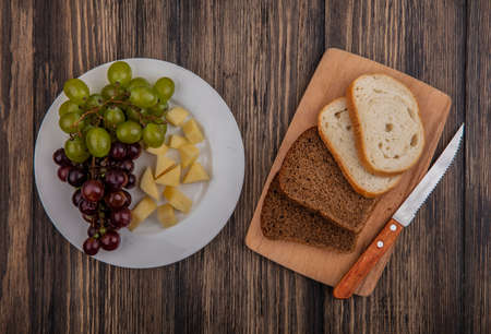 top view of breads as sliced rye and white ones with knife on cutting board and plate of grape and cheese on wooden backgroundの写真素材