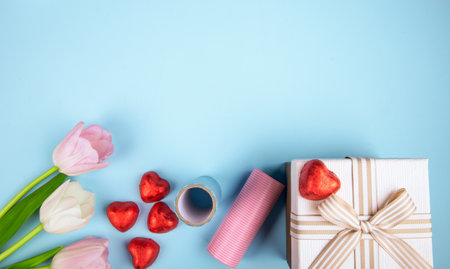 top view of pink color tulips heart shaped chocolate candies wrapped in red foil, gift box and roll of colorful paper on blue background with copy spaceの写真素材