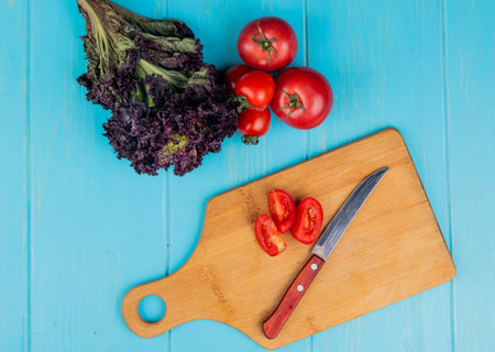 top view of sliced tomatoes with knife on cutting board and basil with whole tomatoes on blue...