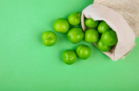 top view of green sour plums scattered from a sack on green background