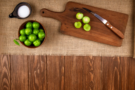 top view of sour green plums in a bowl and sliced plums on a wooden cutting board with knife and salt on sackcloth on dark rustic background with copy spaceの写真素材