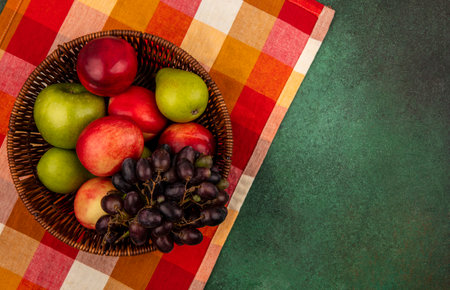 top view of fruits as peach apple pear grape in basket on plaid cloth and on green background with copy spaceの写真素材