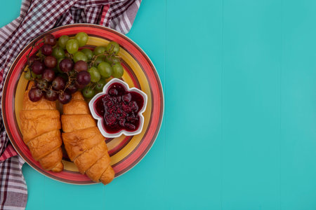 top view of croissants with grapes and raspberry jam in plate on plaid cloth on blue background with copy spaceの写真素材