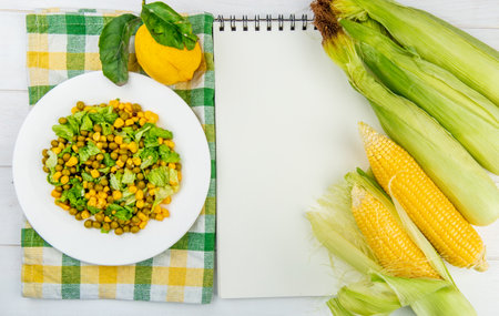 top view of corn salad on cloth and lemon with corn cobs and note pad on wooden backgroundの写真素材