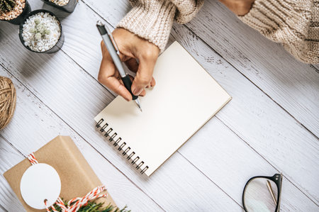 A hand holding a pen to write on a notebook with glasses and a small gift on a white wooden floor