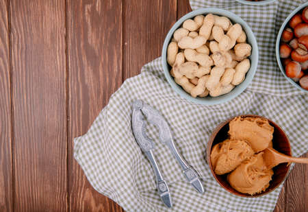 top view of peanut butter in a wooden bowl with hazelnuts and peanuts in shell in bowls and nut...