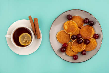 top view of pancakes with cherries in plate and cup of tea with lemon slice in it and cinnamon on...